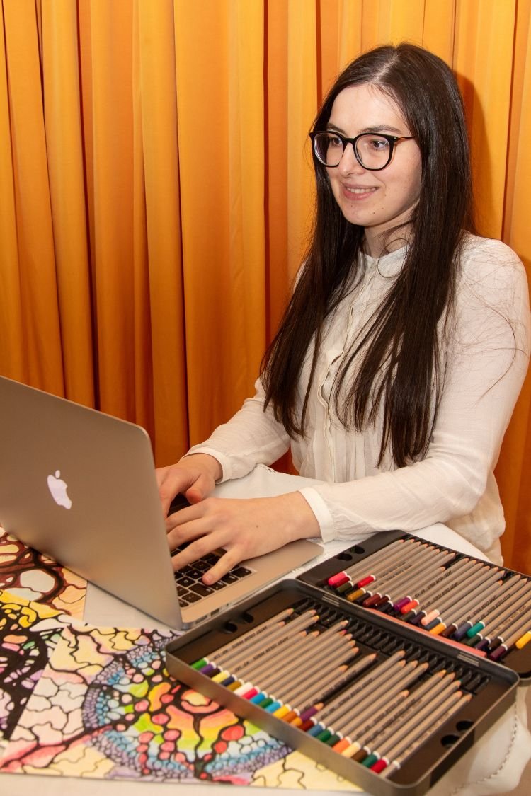 A woman with long dark hair and glasses (Ana Jomir Neurographica Coach and Instructor) smiles while working on a MacBook at a desk covered with colorful Neurographica artwork and an open case of coloured pencils