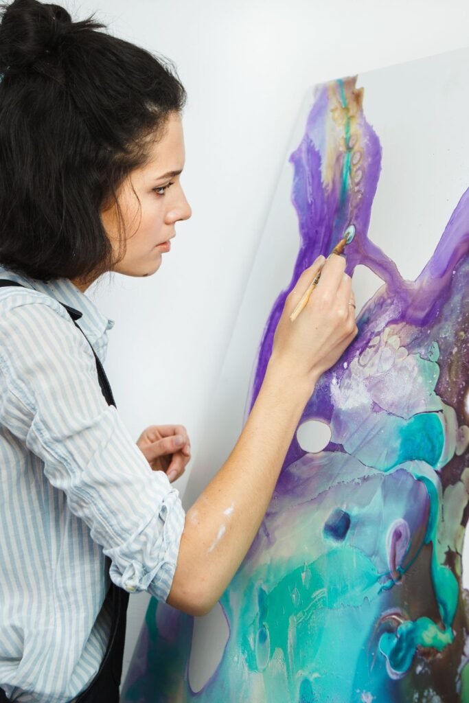 A young woman with dark hair tied up is focused on painting an abstract artwork using vibrant purple, turquoise, and teal hues. She holds a fine brush and is adding detail to an organic, flowing shape on a vertical canvas. She wears a striped shirt and dark overalls, suggesting a creative or therapeutic setting.