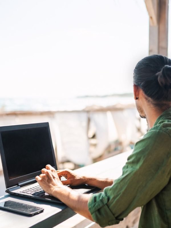 Digital nomad working A man with a tied-back hairstyle wearing a green shirt works on a laptop at an outdoor beachside location, with a smartphone beside him on a wooden table and a view of the sea in the background.